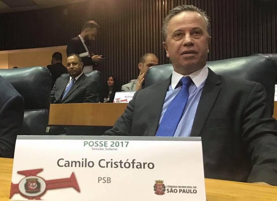 Vereador Camilo Cristófaro sentado à mesa na Câmara Municipal de São paulo. Diante dele, uma placa traz o nome do vereador e o partido "PSB". O parlamentar tem pele clara, entradas no cabelo sobre a testa e os fios do cabelo são grisalhos.