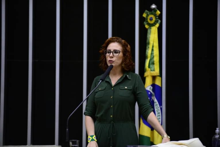 Carla Zambelli discursa no púlpito da câmara dos Deputados. Atrás dela há uma bandeira.